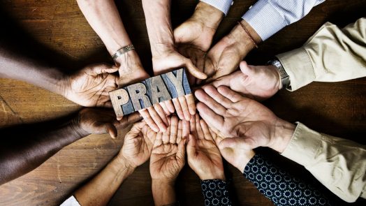 Many hands grabbing a woodblock that spells Pray