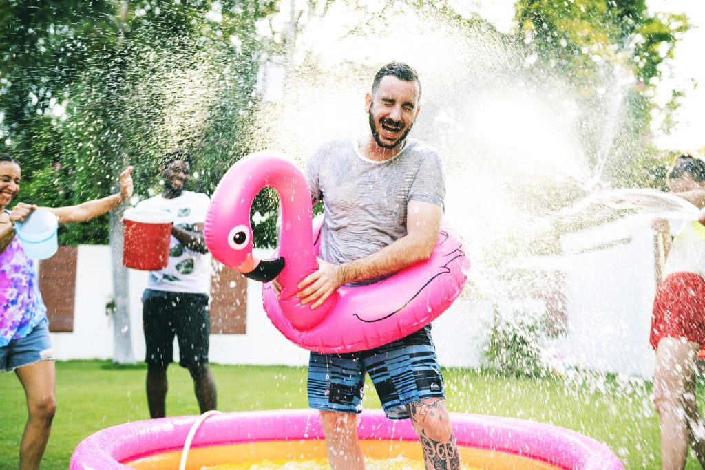 Man in a kid pool getting sprayed with water by his family and friends
