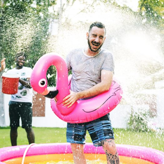 Man in a kid pool getting sprayed with water by his family and friends